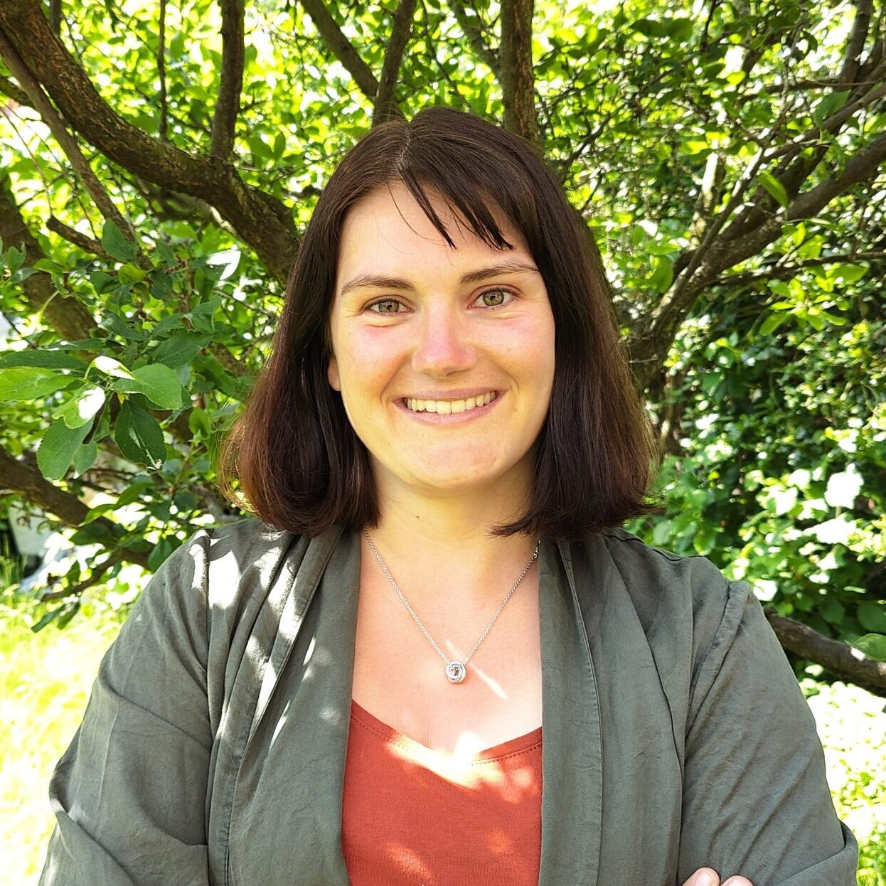 Profile photo of Charlotte Flechet, smiling with her arms crossed, standing under a tree